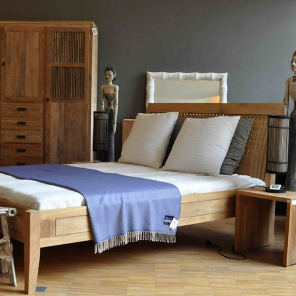 Massivholz Bett Siena aus Teak im klassischen Design in der MOEBEL KOLONIE Farbe Roh, Edelholzbett aus Teakholz ein Schreiner Meisterstueck für das Schlafzimmer, Massivholz Teak Schrank im Hintergrund, Kissen und Plaid als Accessoires, Teakholz Moebel Muenchen